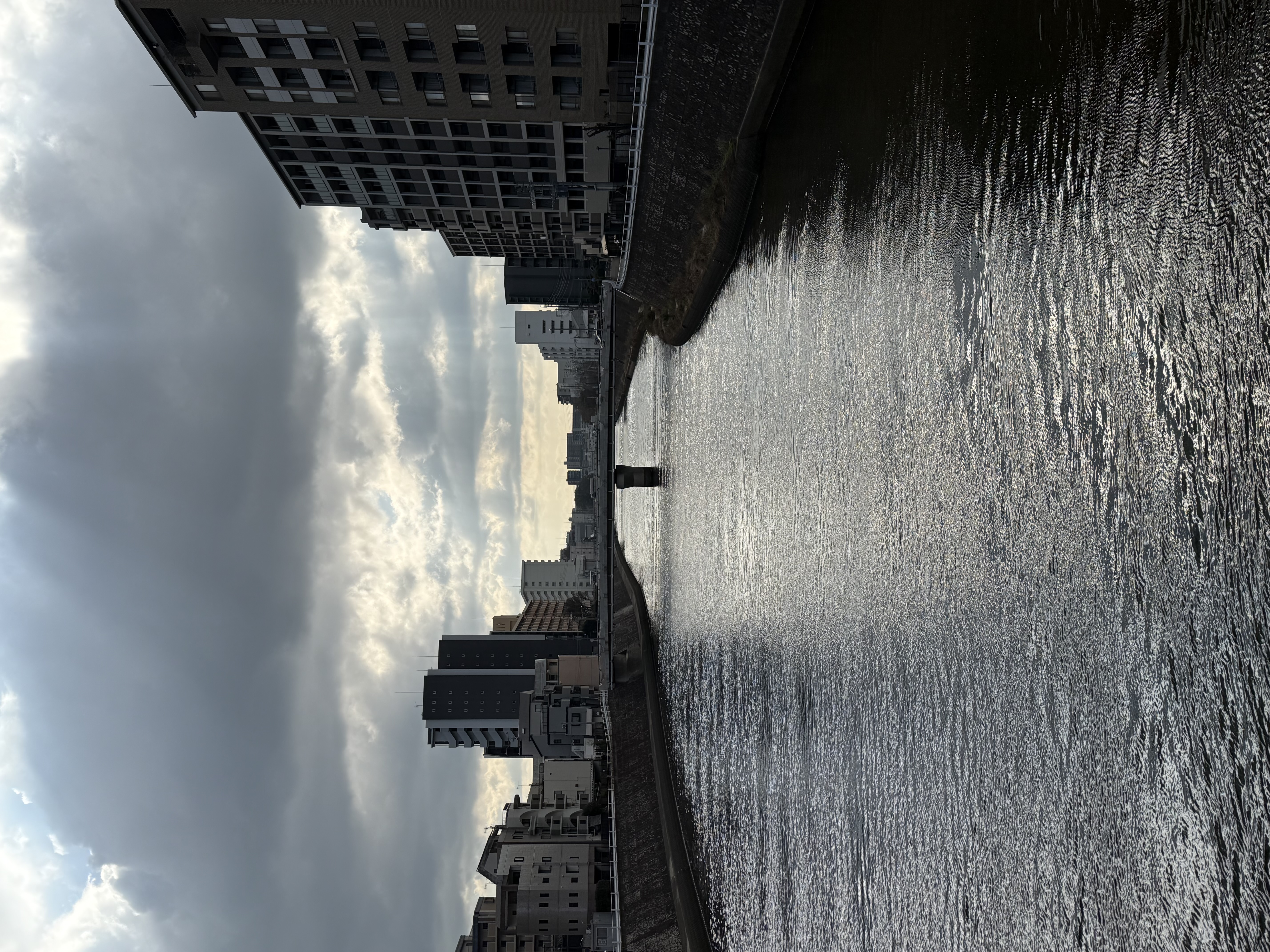 A light breeze makes a wide, calm river slightly choppy, flanked on both sides by concrete embankments and apartment buildings. God rays peek out from