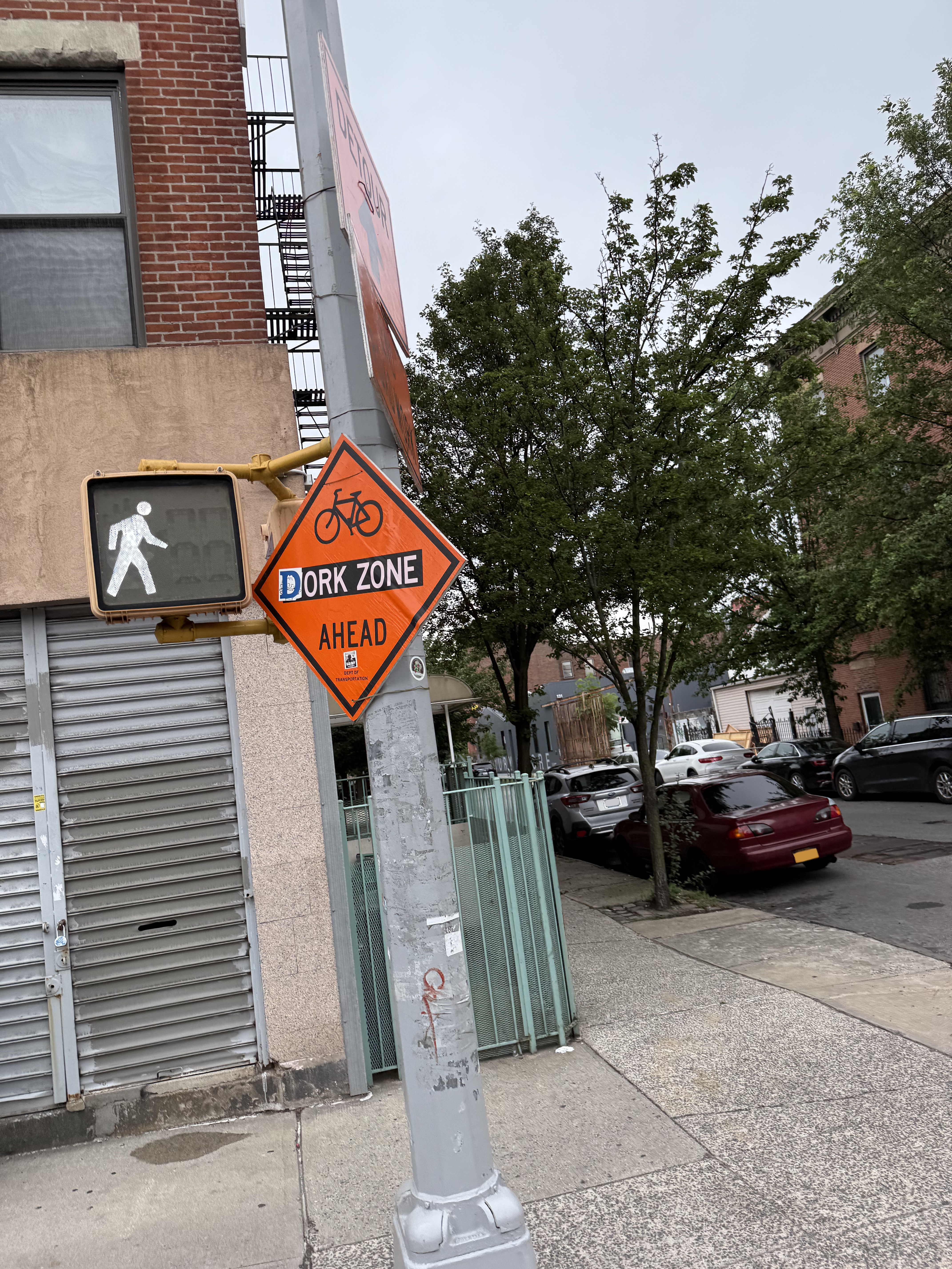 orange diamond sign has been modified to say "Dork zone ahead", photo taken at a dutch angle of an urban car-lined street