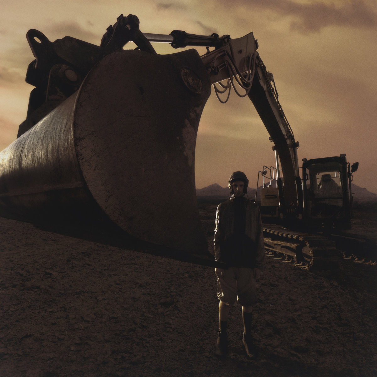 A construction worker stands in front of an excavator, its claw looming over his shoulder. They are in the middle of a desolate wasteland, everything color-graded a muddy brown