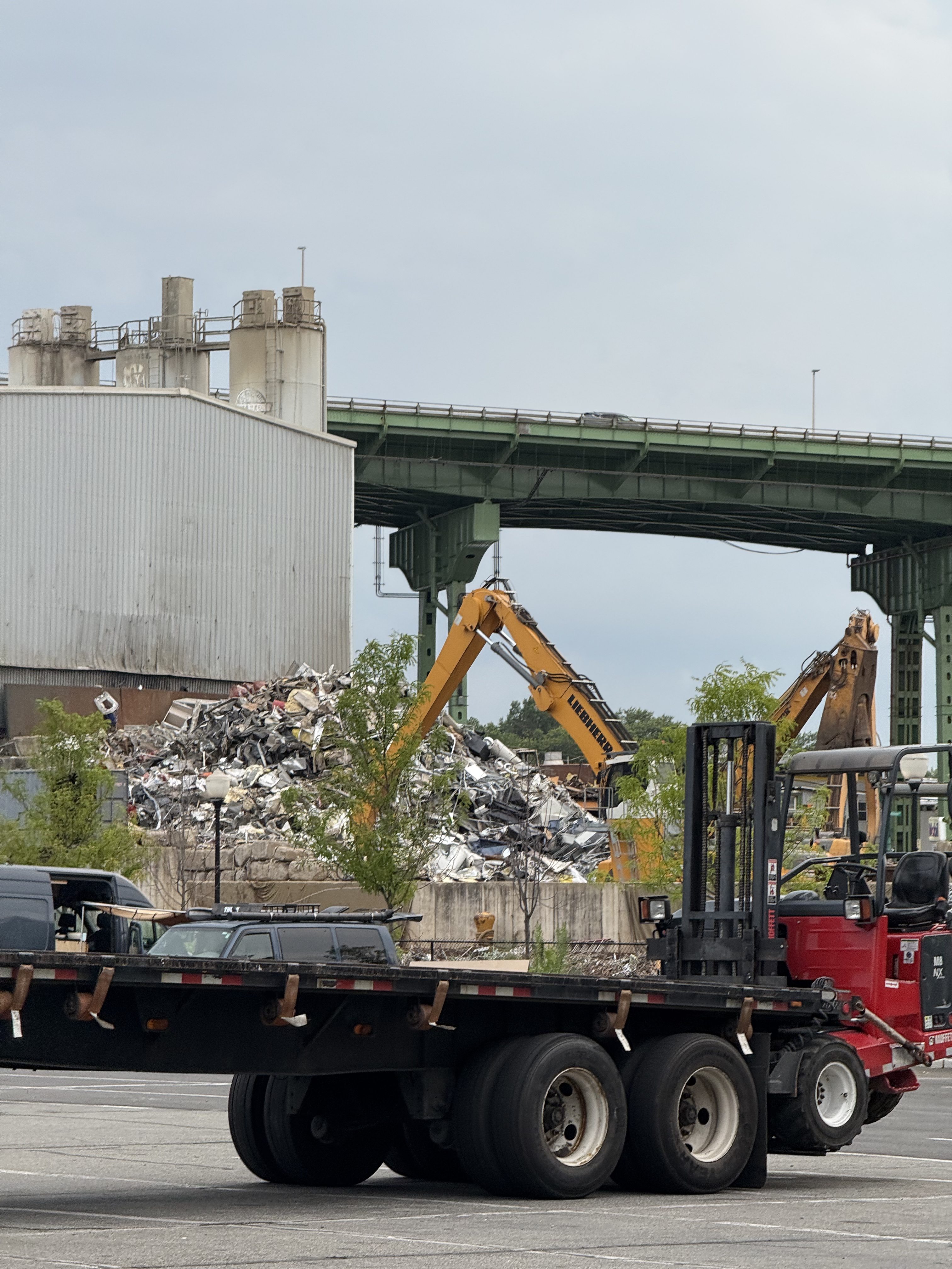 a flatbed in front of a forklift in front of a camper van in front of a garbage dump combed over by excavators in front of an elevated highway