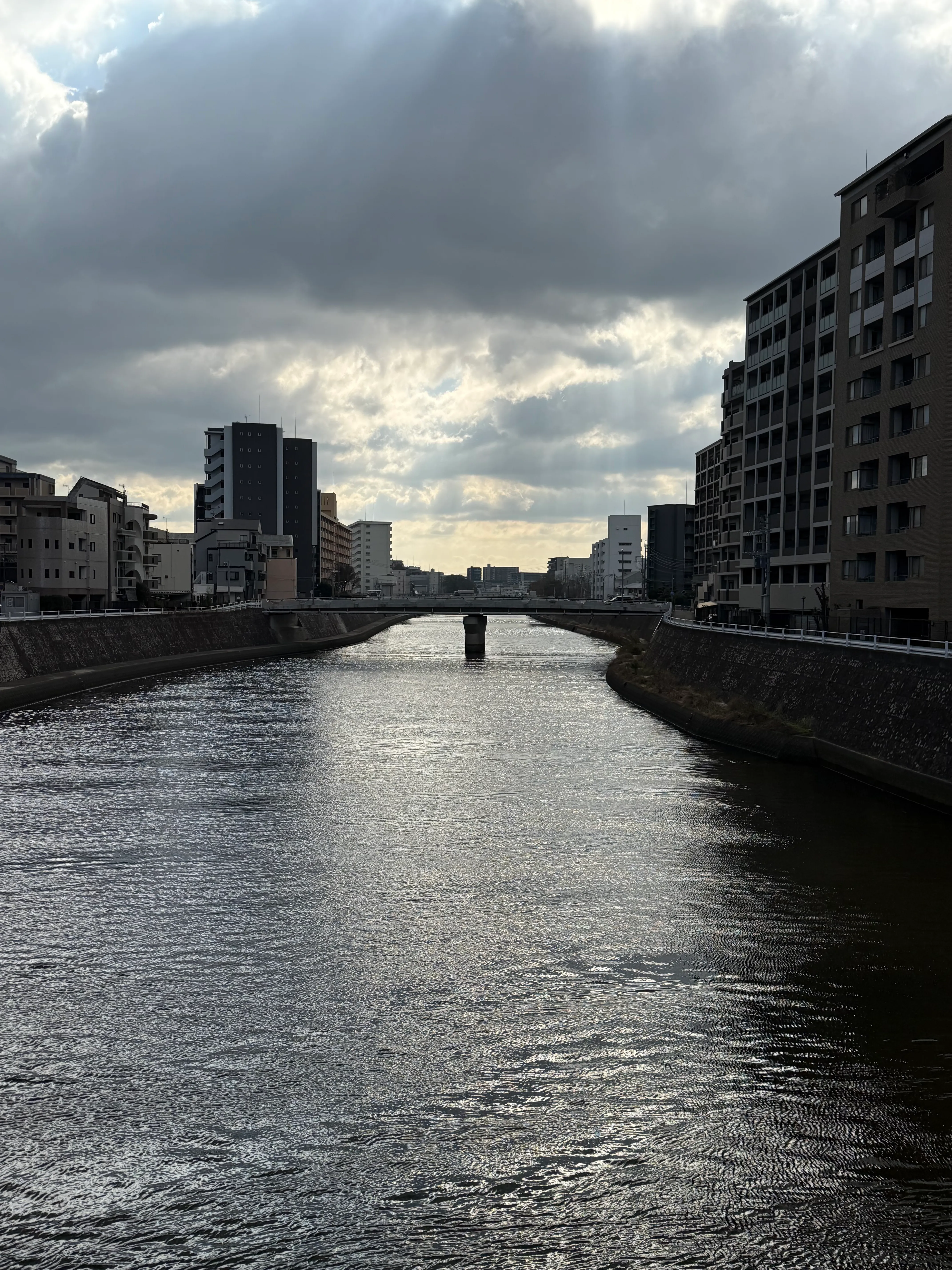 A light breeze makes a wide, calm river slightly choppy, flanked on both sides by concrete embankments and apartment buildings. God rays peek out from