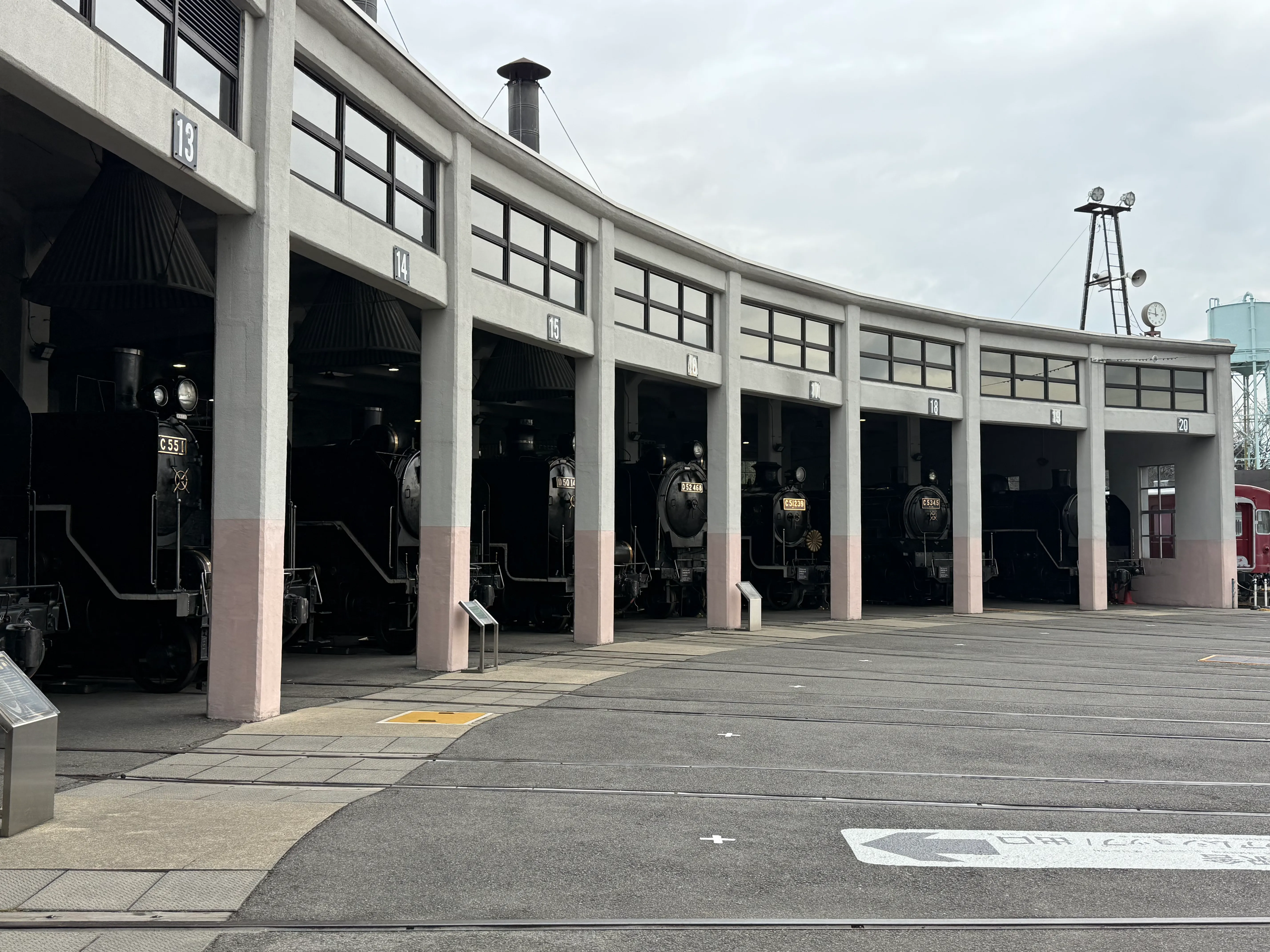 Multiple black steam locomotives are lined up in housing bays in a large arc around a turntable