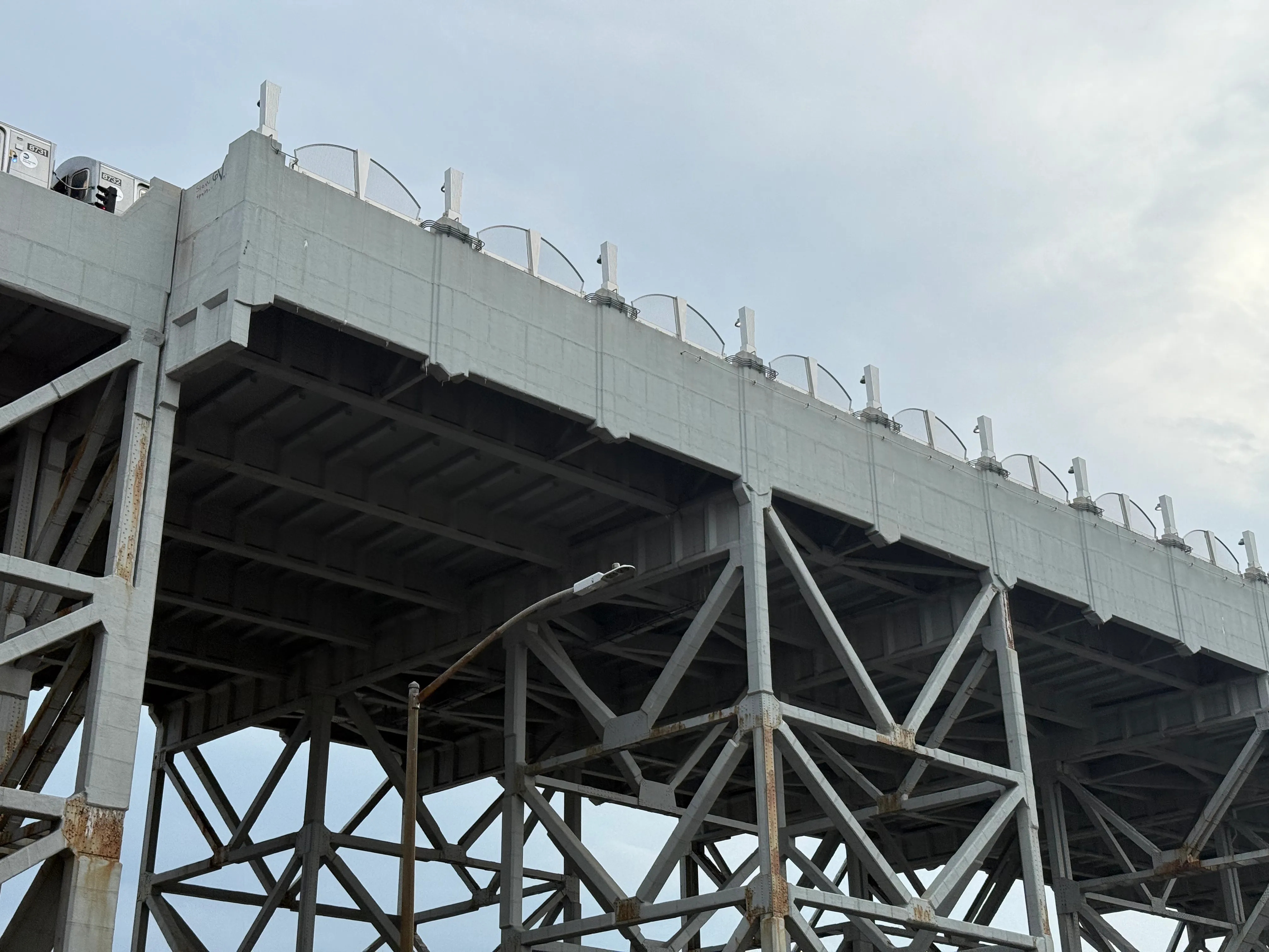 below the train tracks, a large set of steel girders hold up the blue-beige bridge they run on. it&#x27;s almost the same color as the sky